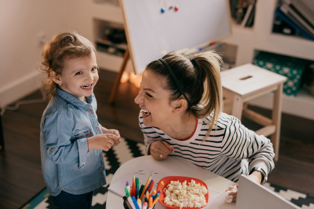 mom with child laughing in arts and crafts area with bowl of popcorn and pens
